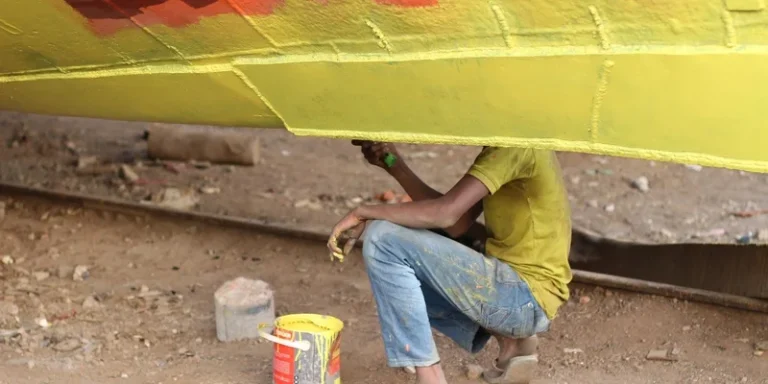 A worker wearing yellow paints a boat's hull in a shipyard