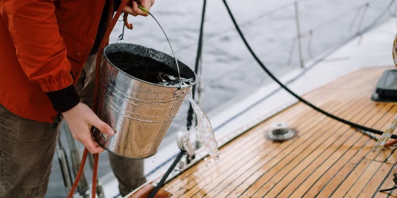 Close-up of a man washing a sailboat deck with water from a metal bucket outdoors