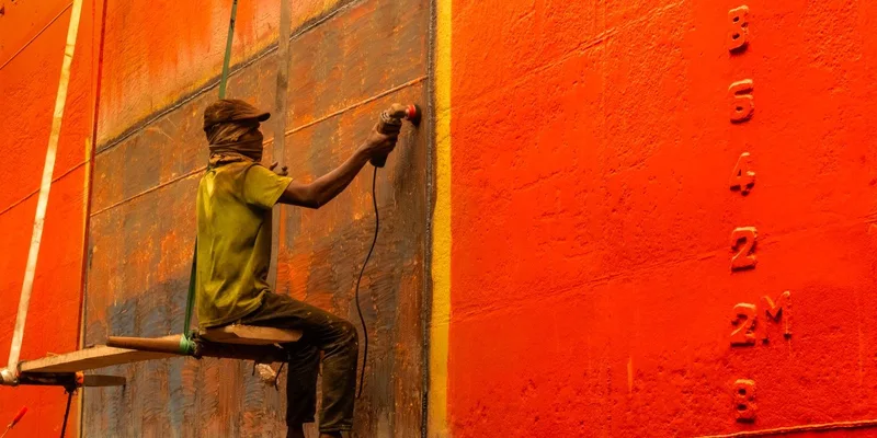 A worker carefully sands the vibrant orange hull of a ship, engaged in maritime maintenance