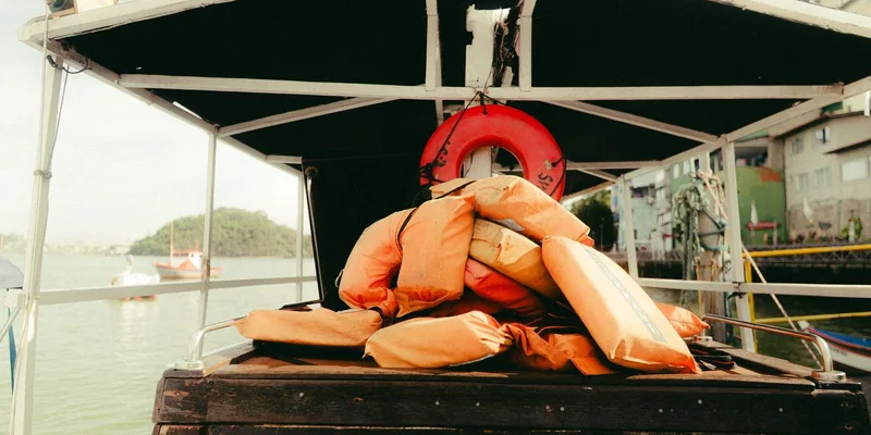 A stack of orange life vests and a life ring on a boat near a calm waterfront