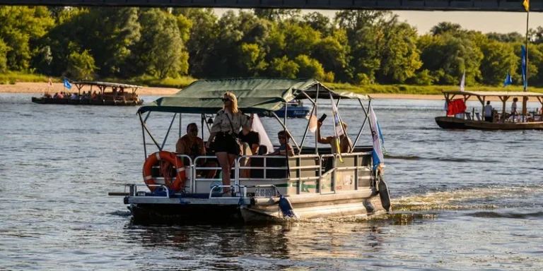 A pontoon boat with people enjoying a sunny day on a river beneath a bridge