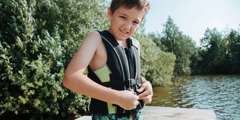 Happy child in a life jacket smiling at the camera while standing on a lakeside jetty