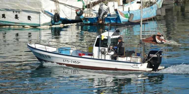 A man navigates a motorboat through a harbor, with colorful fishing boats in the background