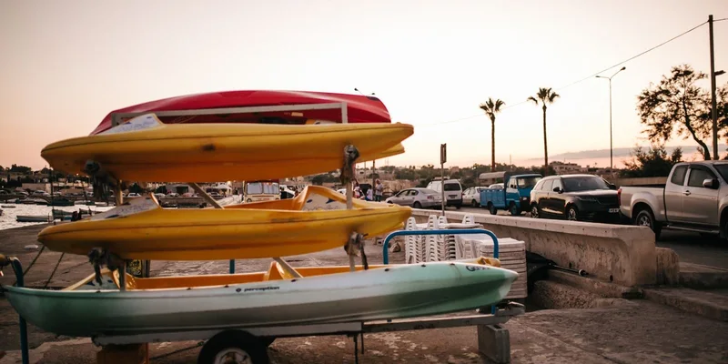 A stack of colorful kayaks at a marina during sunset, with parked cars nearby